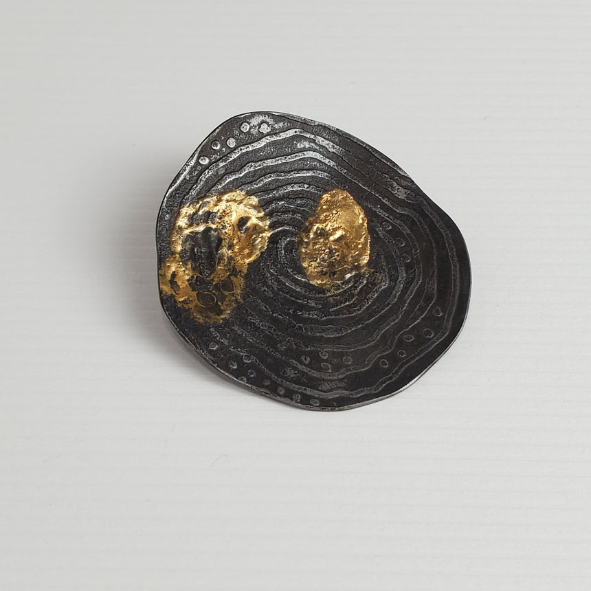 Roundish steel with slightly etched lines and dots that have been patinaed black.  On top of this 24 karat gold has been fused with the steel.  Because this is done with heat, there is little control over how the gold webs and melts in splotches over the steel.  There is a silver tube which can be added to the pin back and then a cord, chain or neckwire can be threaded through the tube to wear as a pendant.
