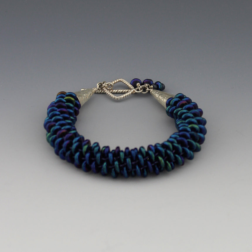 This lovely peacock colored bracelet is made up of off center Czech lentil beads which are woven in a kumihimo braid. Kumihimo is a Japanese weave of 8 different strands on a marudai. The ends are capped with hammered sterling silver cones and a  square twisted wire clasp. Tiny sapphire beads are used to create a dangle.