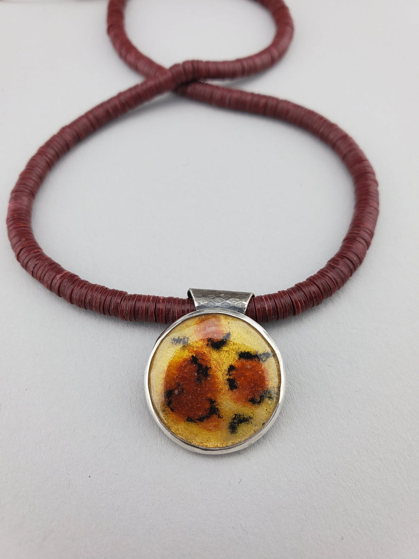 A round pendant with a golden background, the brown areas and the black markings of a leopard print.  Mounted in a silver bezel with a textured backplate and threaded onto a necklace of rust colored African record beads.  Sterling silver lobster clasp.
