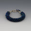 This lovely peacock colored bracelet is made up of off center Czech lentil beads which are woven in a kumihimo braid. Kumihimo is a Japanese weave of 8 different strands on a marudai. The ends are capped with hammered sterling silver cones and a  square twisted wire clasp. Tiny sapphire beads are used to create a dangle.