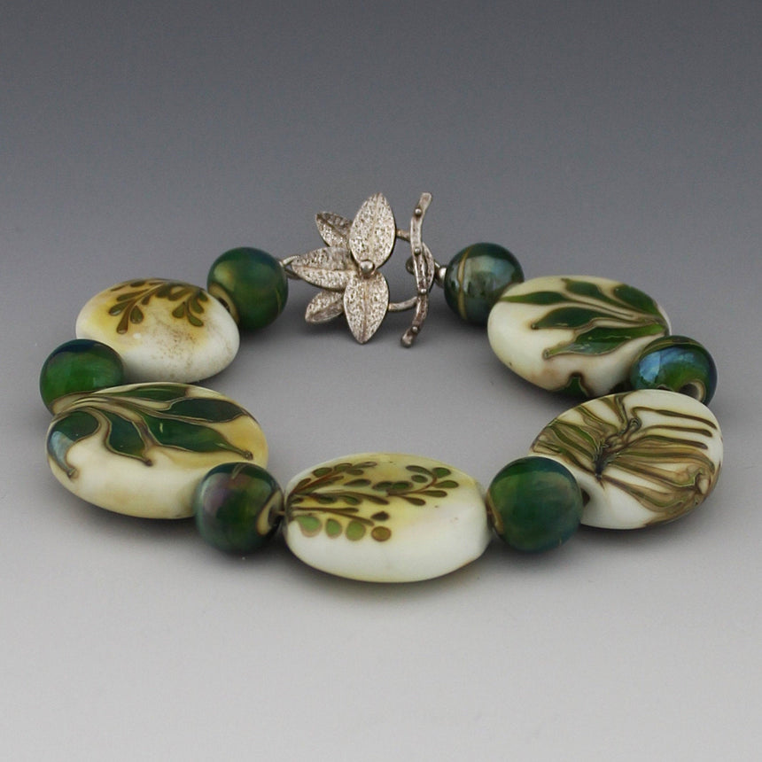 Five organic lentil beads in ivory colored glass have the design of leaves on them.  These leaves are inspired by my garden and reflect the fine leafed ferns, palms and broad leafed ginger.  the green glass has a special silver reaction so that is shimmers in blue and gold.  The clasp has five leaves with an "s" catch.