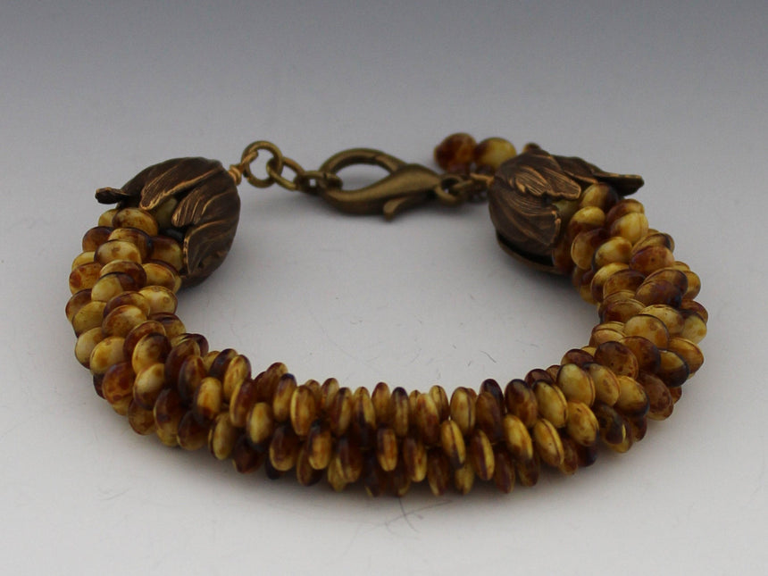 This lovely bracelet is made up of off center Czech lentil beads in tortoise shell colors of brown and gold; which are woven in a kumihimo braid.  Kumihimo is a Japanese weave of 8 different strands on a marudai.  The ends are capped with bronze  acanthus cones and a bronze lobster clasp.  Tiny tiger eye beads are used to create a dangle.
