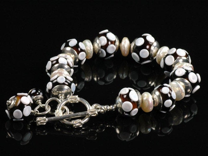 Nine dark brown glass beads have raised white dots.  Each bead is capped on both sides with a silver cap.   Between each silver cap is a cream flat lentil pearl.  A sterling silver toggle clasp with an additional glass bead dangle finishes off this bracelet.